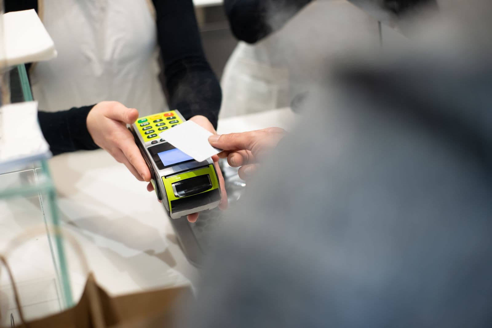A customer holds a plain white card over a handheld payment terminal that a shop assistant is holding out across a counter.