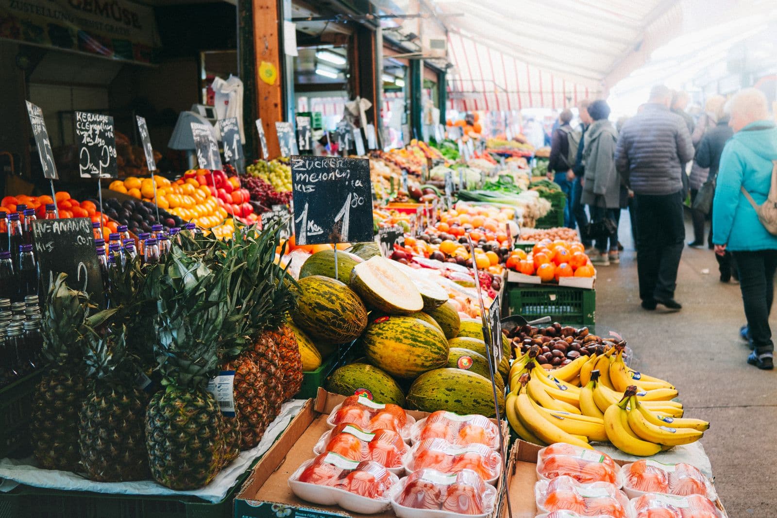Fruit stalls under a striped awning at a Vienna market, with pineapples, melons and bananas in front of handwritten chalkboard price signs and shoppers walking down the aisle.