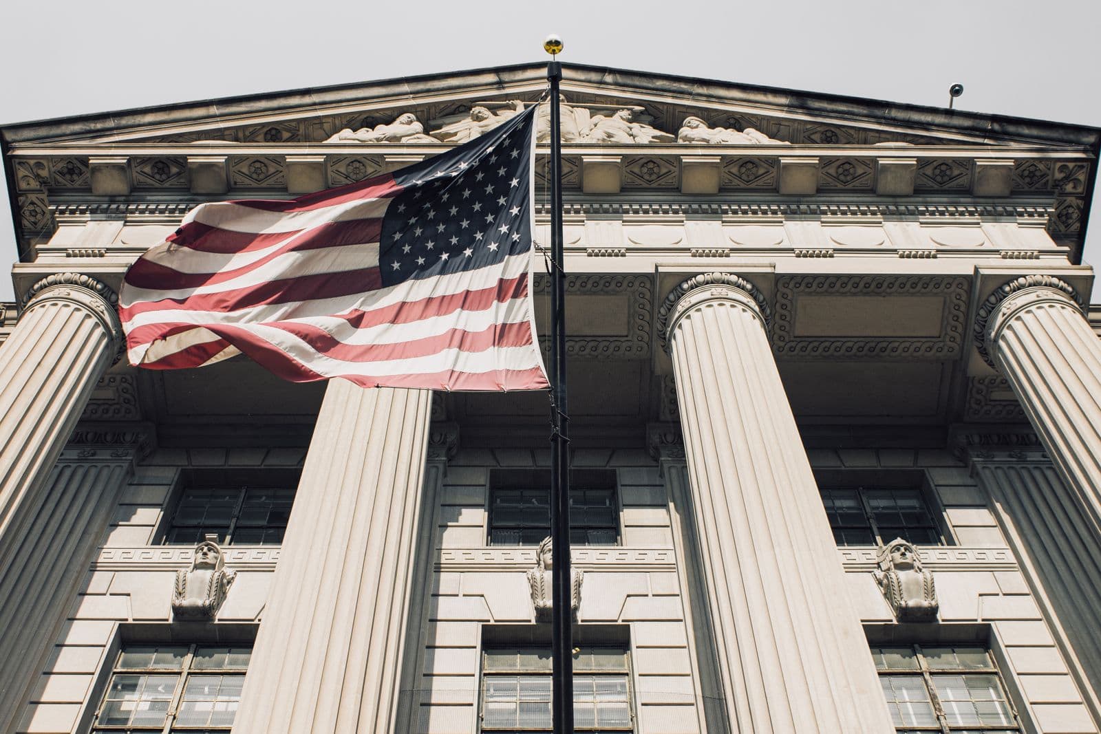 The columned stone front of a federal government building, photographed from below, with a United States flag flying from a pole in front of it.