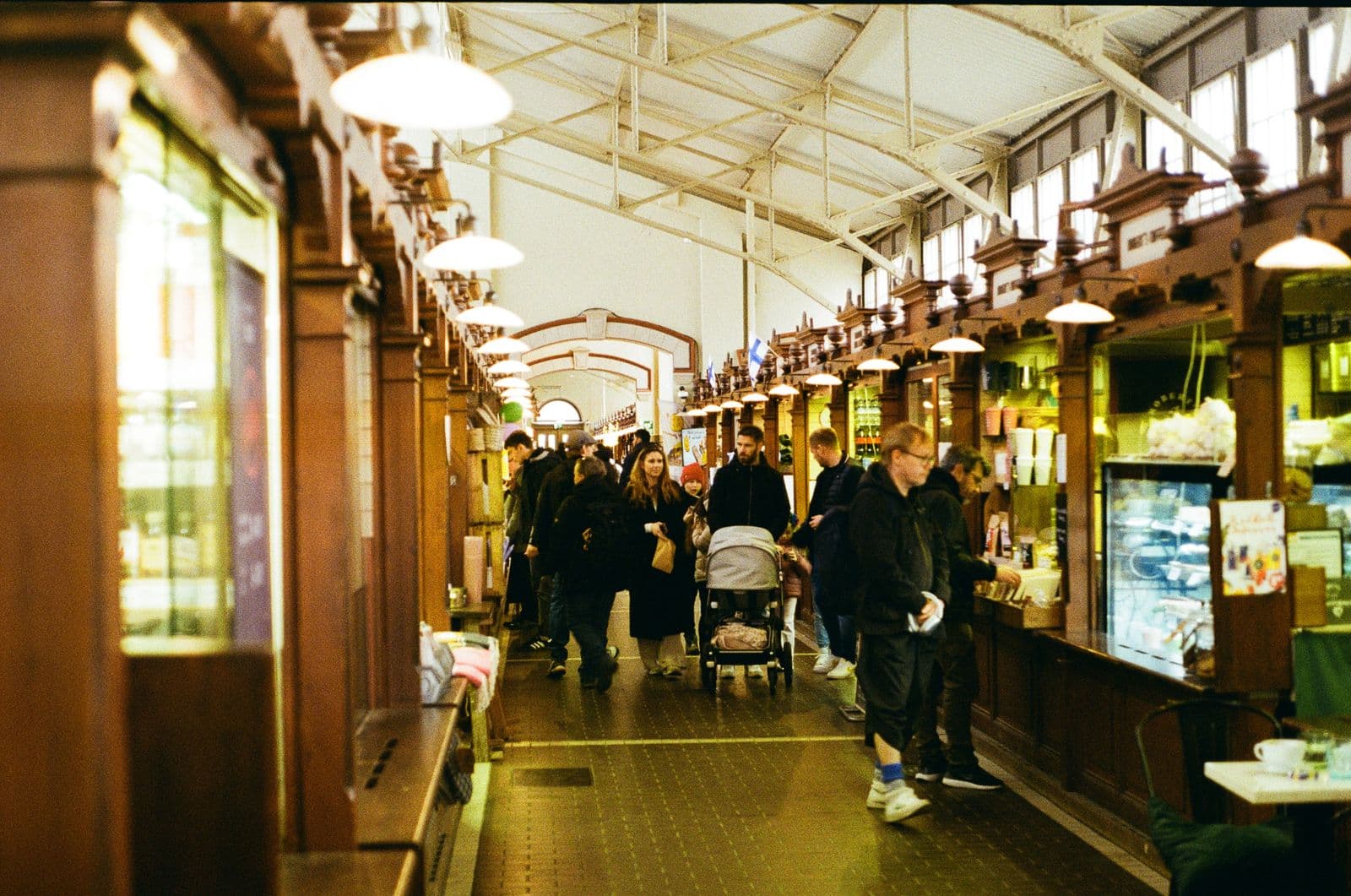 Shoppers, one pushing a pram, walk down the aisle of a historic indoor market hall in Helsinki, between wooden stall fronts lit by hanging lamps, with small Finnish flags above the stalls.