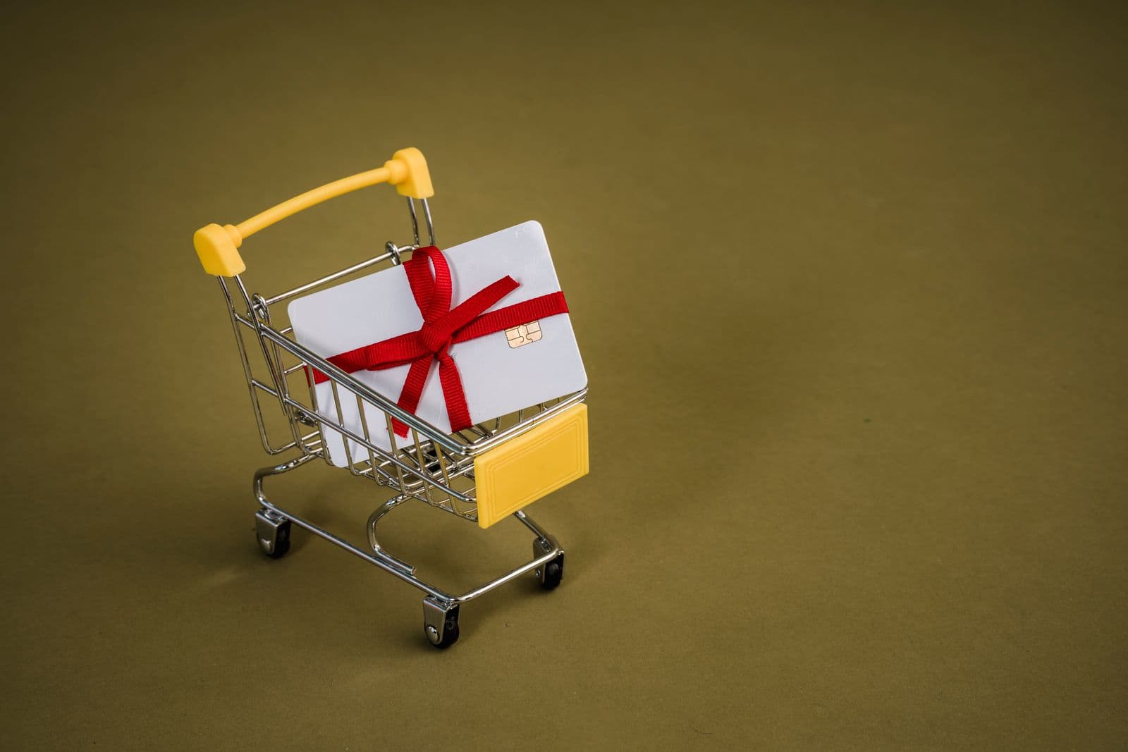 A plain white chip card tied with a red ribbon bow, sitting in a miniature metal shopping cart with yellow handles on an olive-green background.