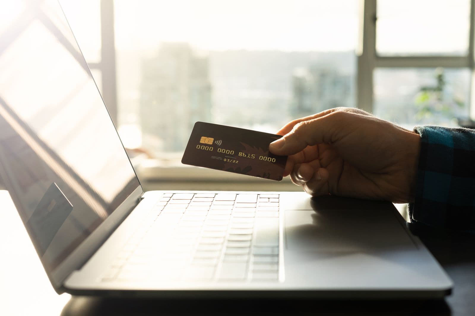 A hand holds a bank card printed with a dummy card number beside an open laptop on a desk, backlit by a bright window.