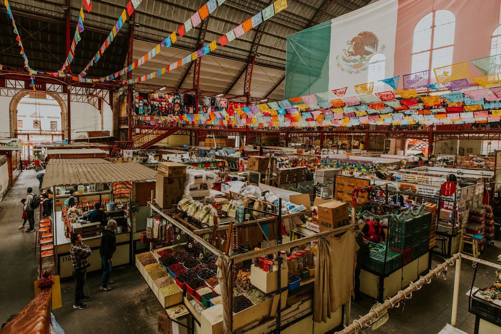 The covered hall of Mercado Hidalgo in Guanajuato seen from a balcony, with rows of small stalls selling dried chillies, spices and snacks under strings of papel picado and a large Mexican flag.