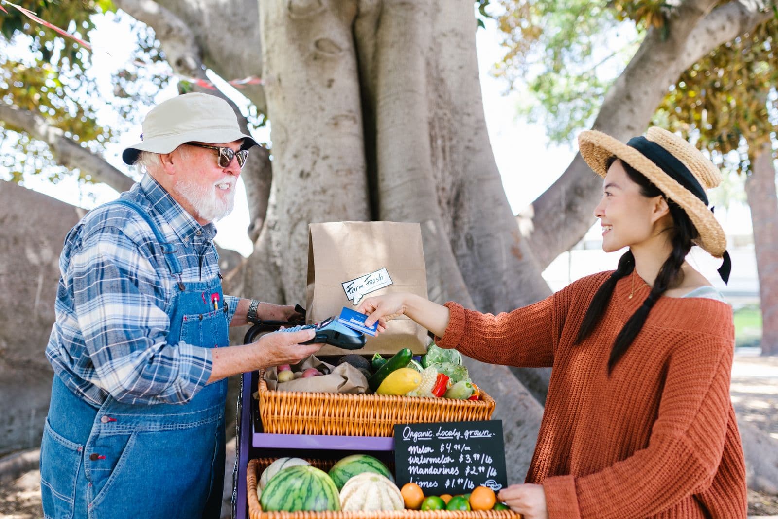 An older man in denim overalls and a sun hat holds a handheld card machine at his fruit and vegetable stall while a smiling woman in a straw hat taps a blue bank card on it, beside a chalkboard of prices.