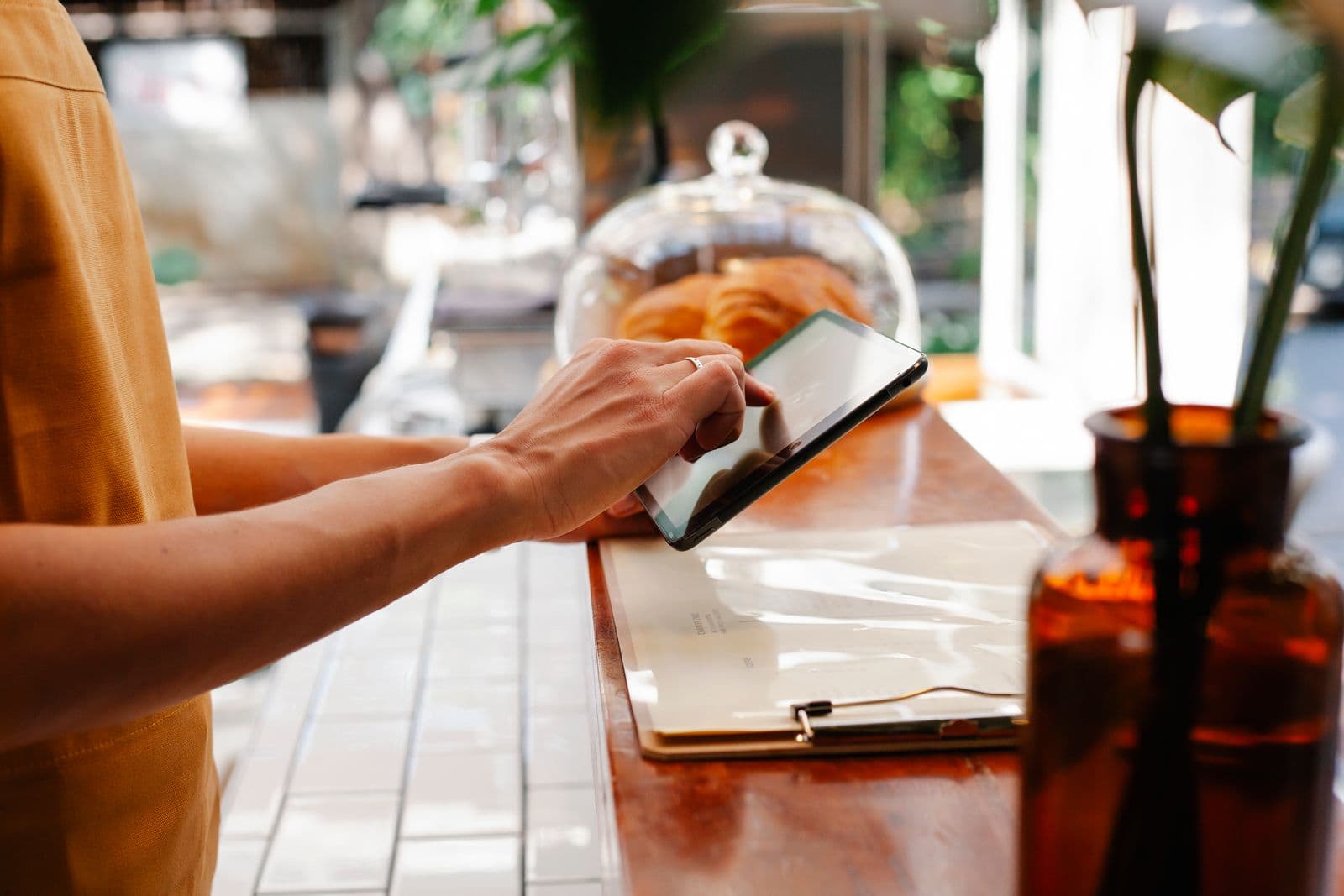 A café worker in a mustard apron leans on a wooden counter and taps a tablet with one finger, beside a clipboard, a glass cake dome with croissants and an amber bottle holding a plant.