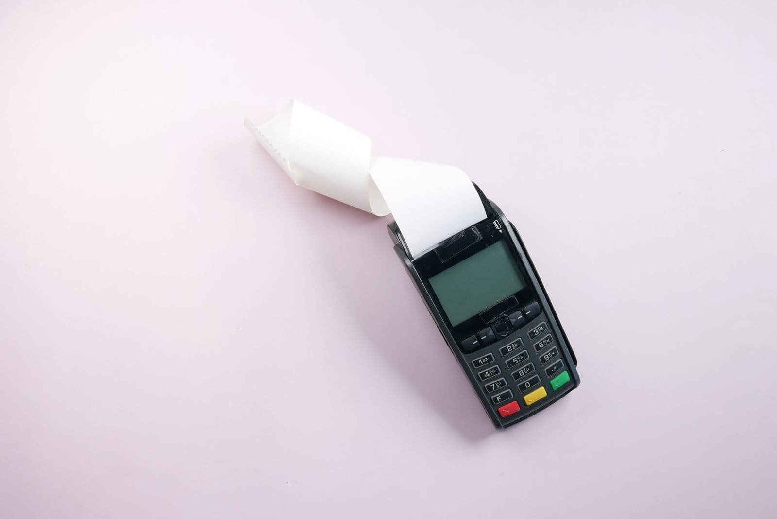 A black card terminal lies on a pale pink surface with a long blank receipt curling out of its printer.