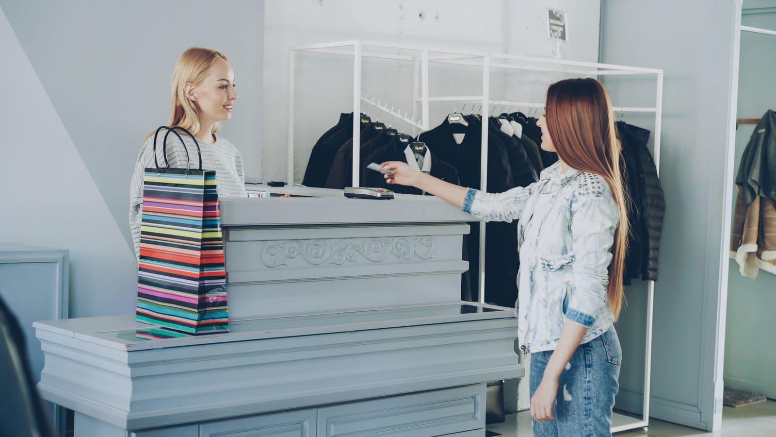In a bright clothing boutique, a customer in a denim jacket holds out a bank card toward the card terminal on the counter, where a smiling shop assistant stands beside a striped shopping bag, with a rail of dark coats behind them.
