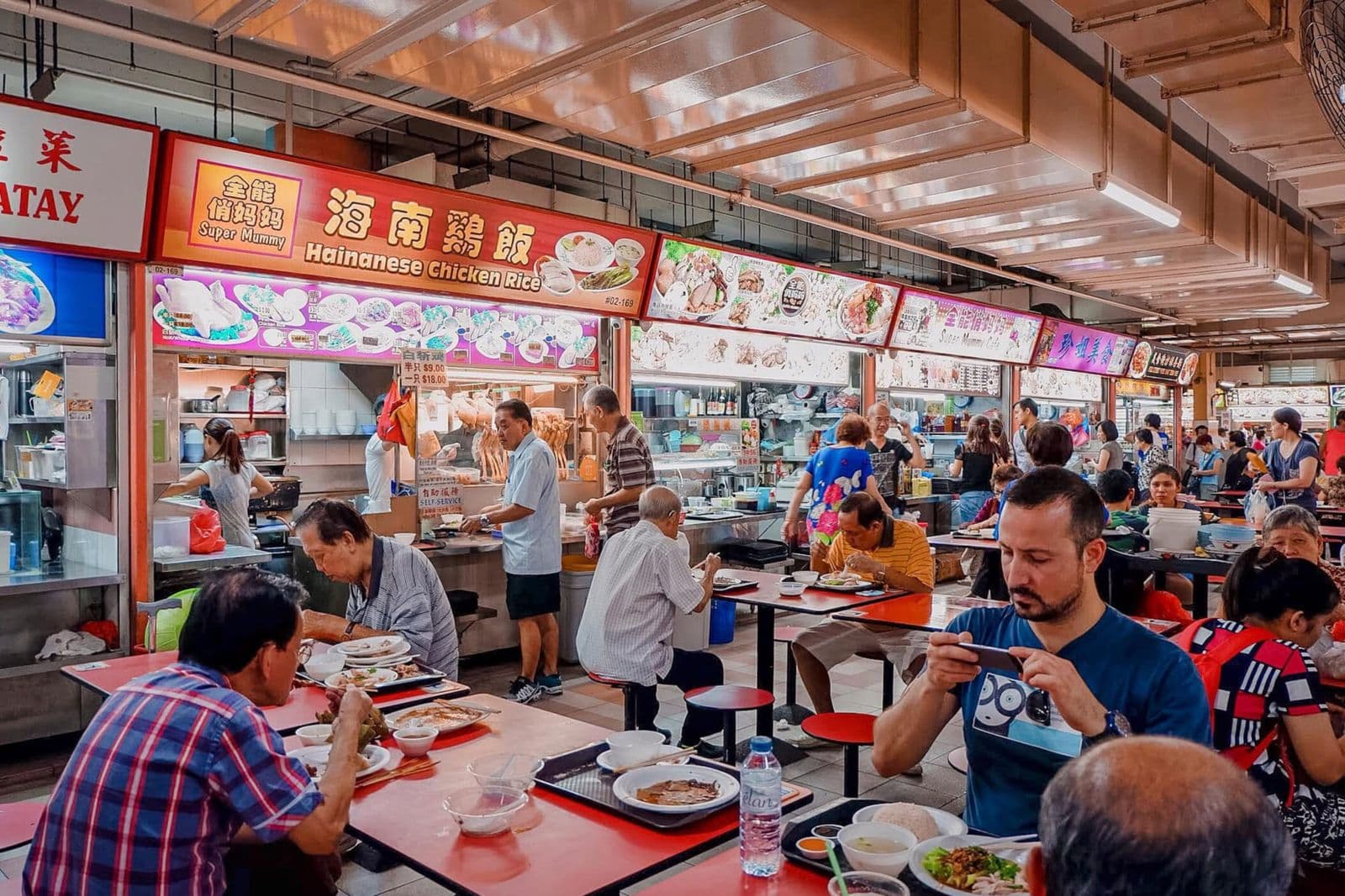 Diners eat at red tables in a busy Singapore hawker centre in front of a row of food stalls, one advertising Hainanese chicken rice, while a man at the front photographs his meal with his phone.
