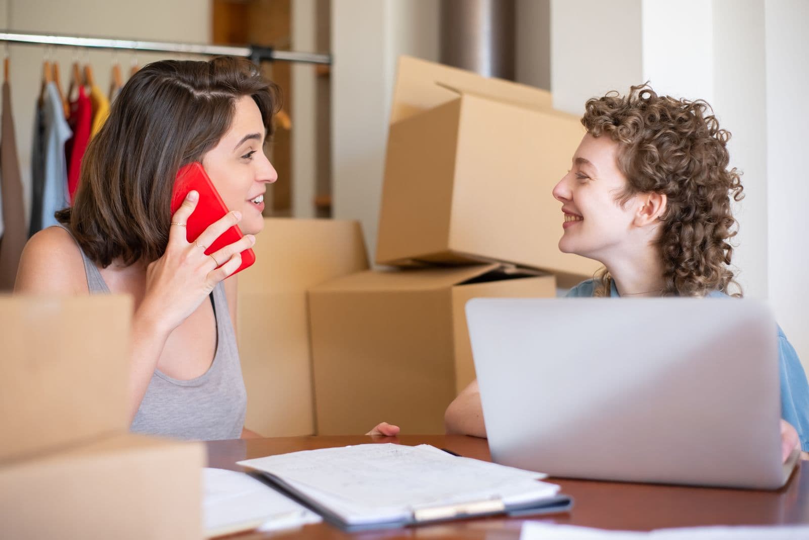 Two women run a small online clothing shop from a table with a laptop and a clipboard, one on the phone, with cardboard boxes and a rail of clothes behind them.
