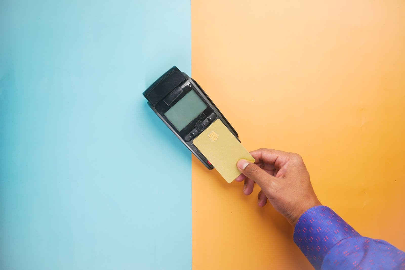 A hand holds a plain gold bank card against the top of a black payment terminal lying on a surface split between pale blue and orange paper.