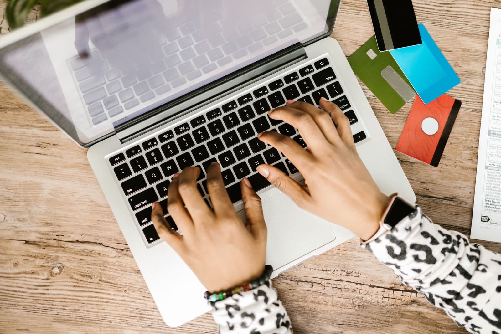 Two hands typing on a laptop keyboard next to four plain payment cards and a printed application form on a wooden desk.