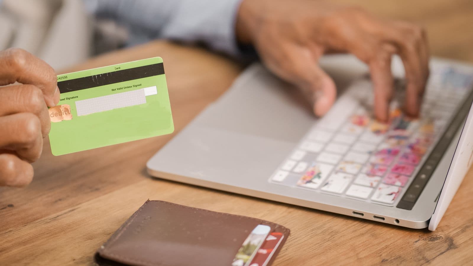 A person holds a plain green payment card in one hand and types its number into a laptop with the other, a leather card wallet lying open on the wooden table beside them.