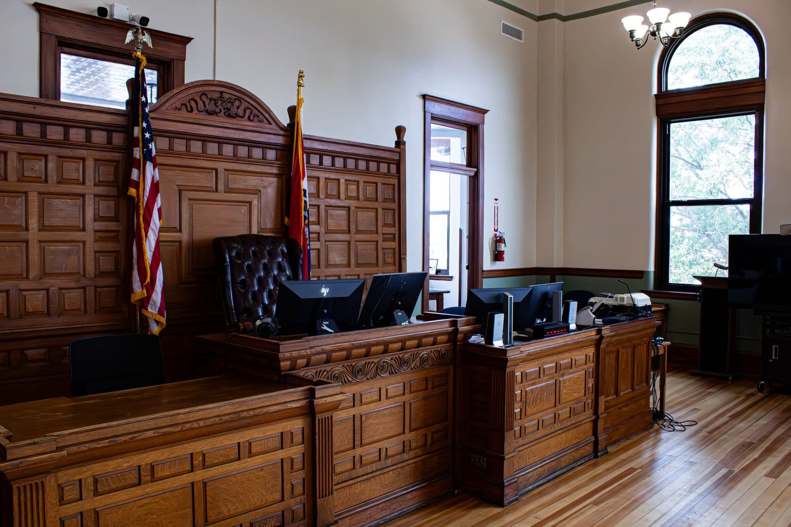 An empty American courtroom with a wood-panelled judge's bench, two flags on poles either side of the leather chair, computer monitors on the clerk's desk and light coming through a tall arched window.