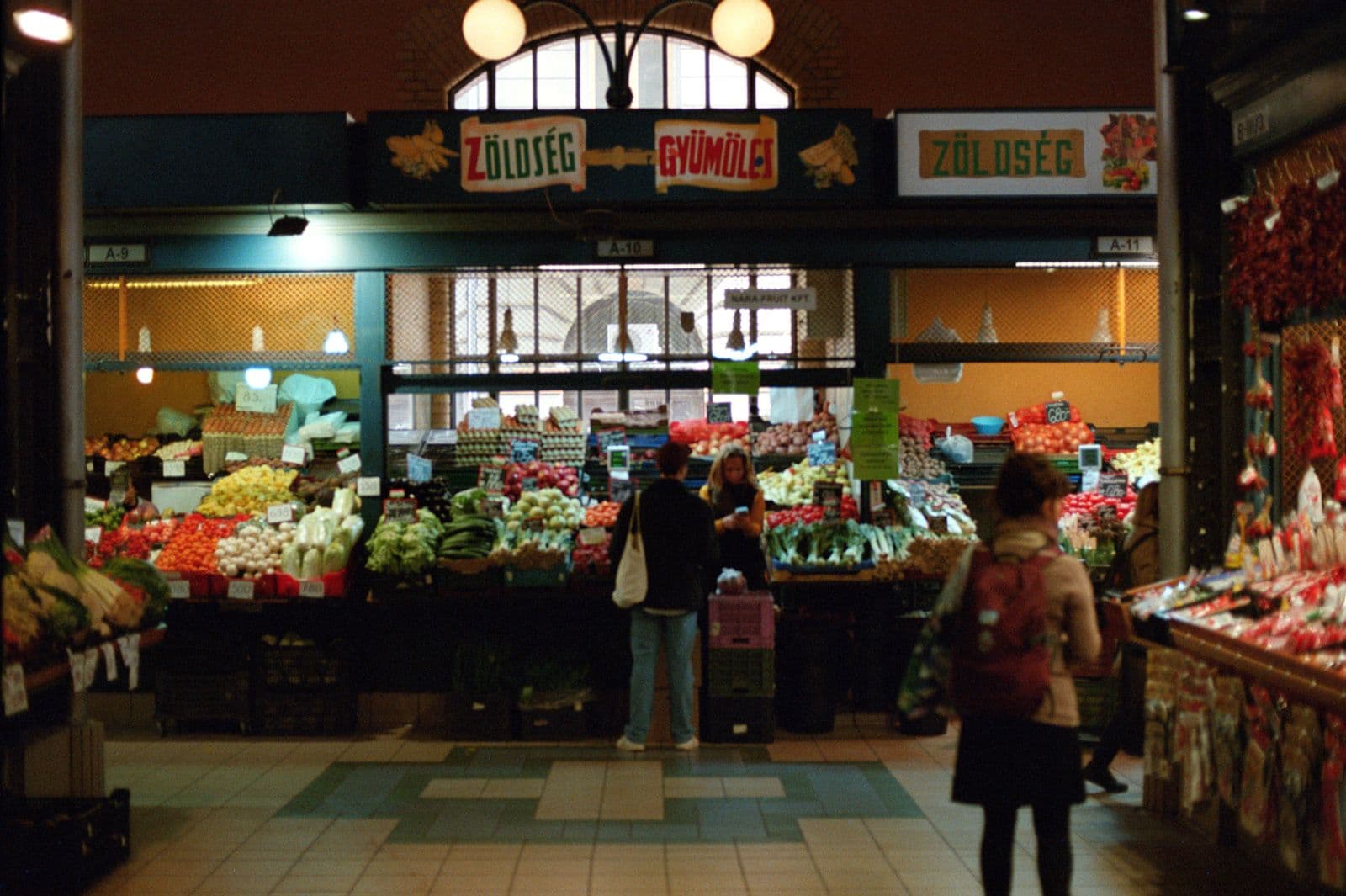 A fruit and vegetable stall inside Budapest's Central Market Hall under painted signs reading Zöldség and Gyümölcs, with a customer at the counter and a shopper with a backpack walking past.