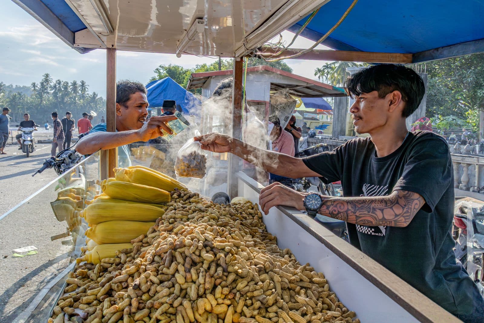 A street vendor at a roadside stall piled with boiled peanuts and corn hands a bag of peanuts to a customer, who holds out a banknote to pay, with palm trees and motorbikes behind them.