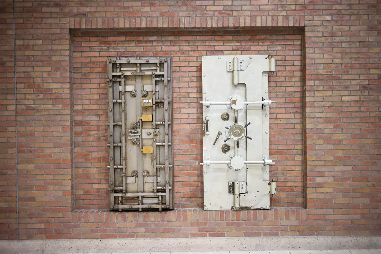 Two heavy steel vault doors mounted side by side in a recess in a red brick wall, one an open framework of gears and bolts, the other a solid plate with a spoked handle.