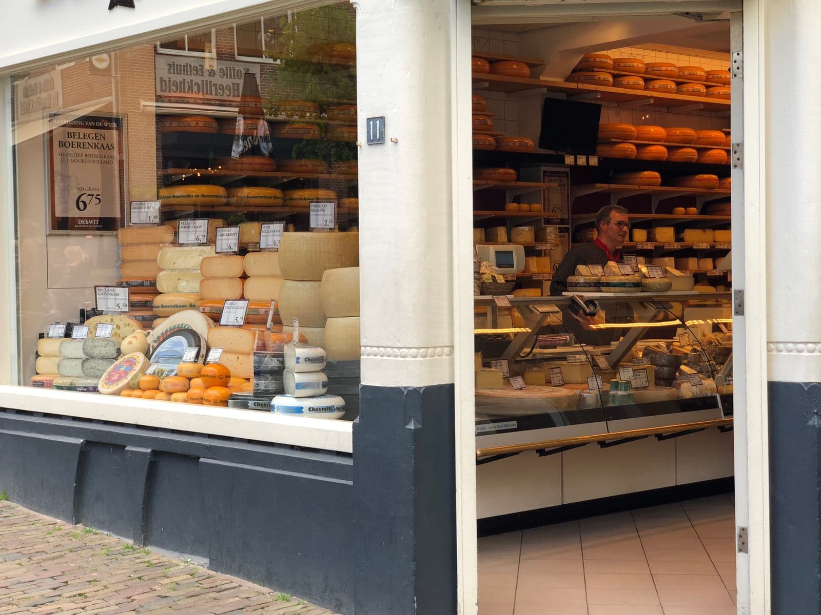 Through the open door of a Dutch cheese shop, a shopkeeper behind the counter holds a card payment terminal, with wheels of Gouda stacked on wooden shelves and priced cheeses in the window.