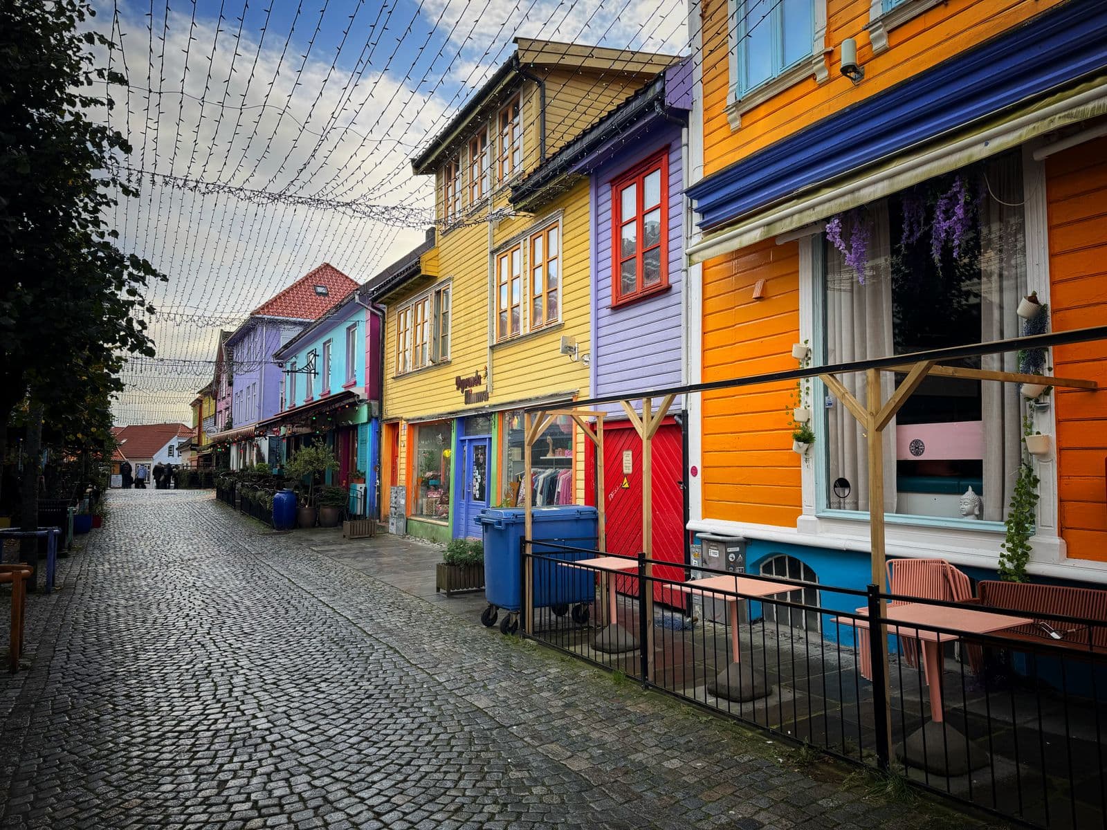 A cobbled shopping street in Stavanger, Norway, lined with brightly painted wooden buildings in yellow, purple and orange, with small shops at street level and strings of lights overhead.