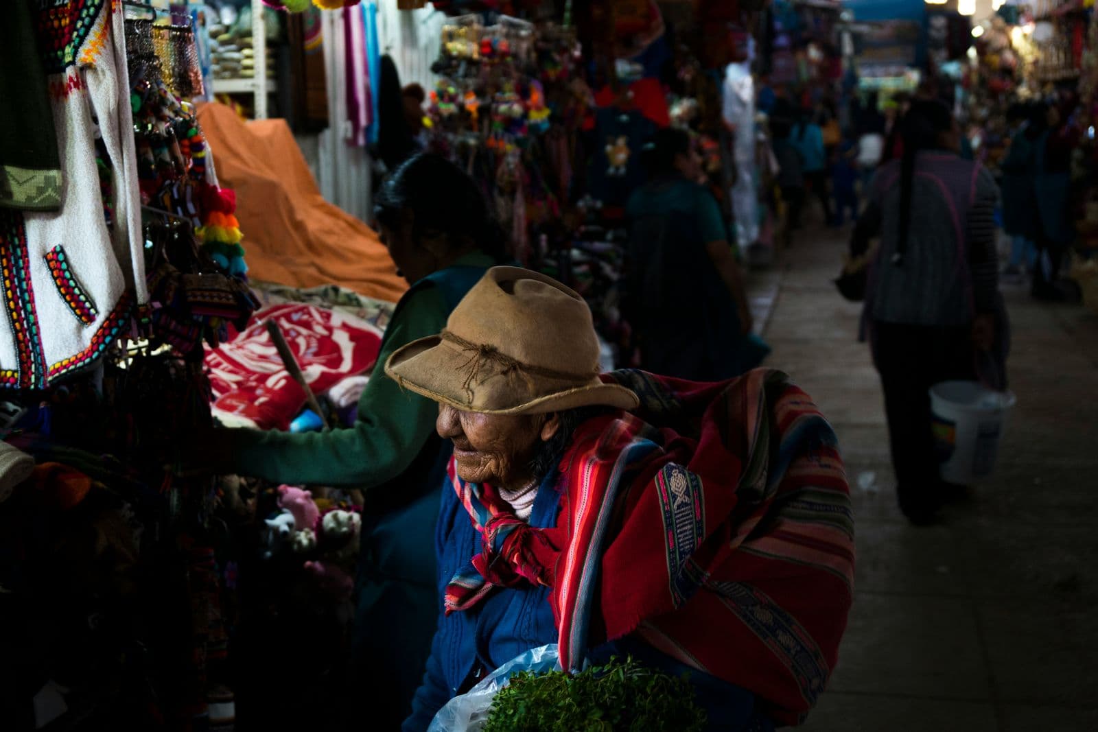An elderly woman in a brown felt hat and a striped red shawl carries a bag of fresh herbs past stalls of woven textiles and knitted goods in a covered Peruvian market, while other shoppers walk down the aisle behind her.