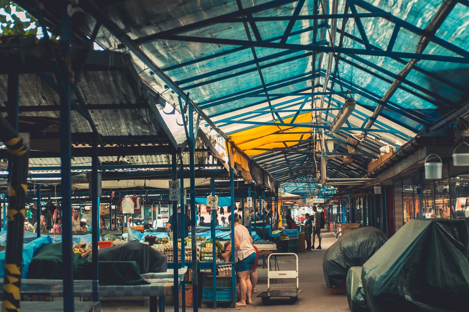Shoppers at fruit and vegetable stalls under the blue steel frame and translucent roof of a covered market in Kraków, Poland, with numbered stall plates and tarpaulin-covered tables along the aisle.