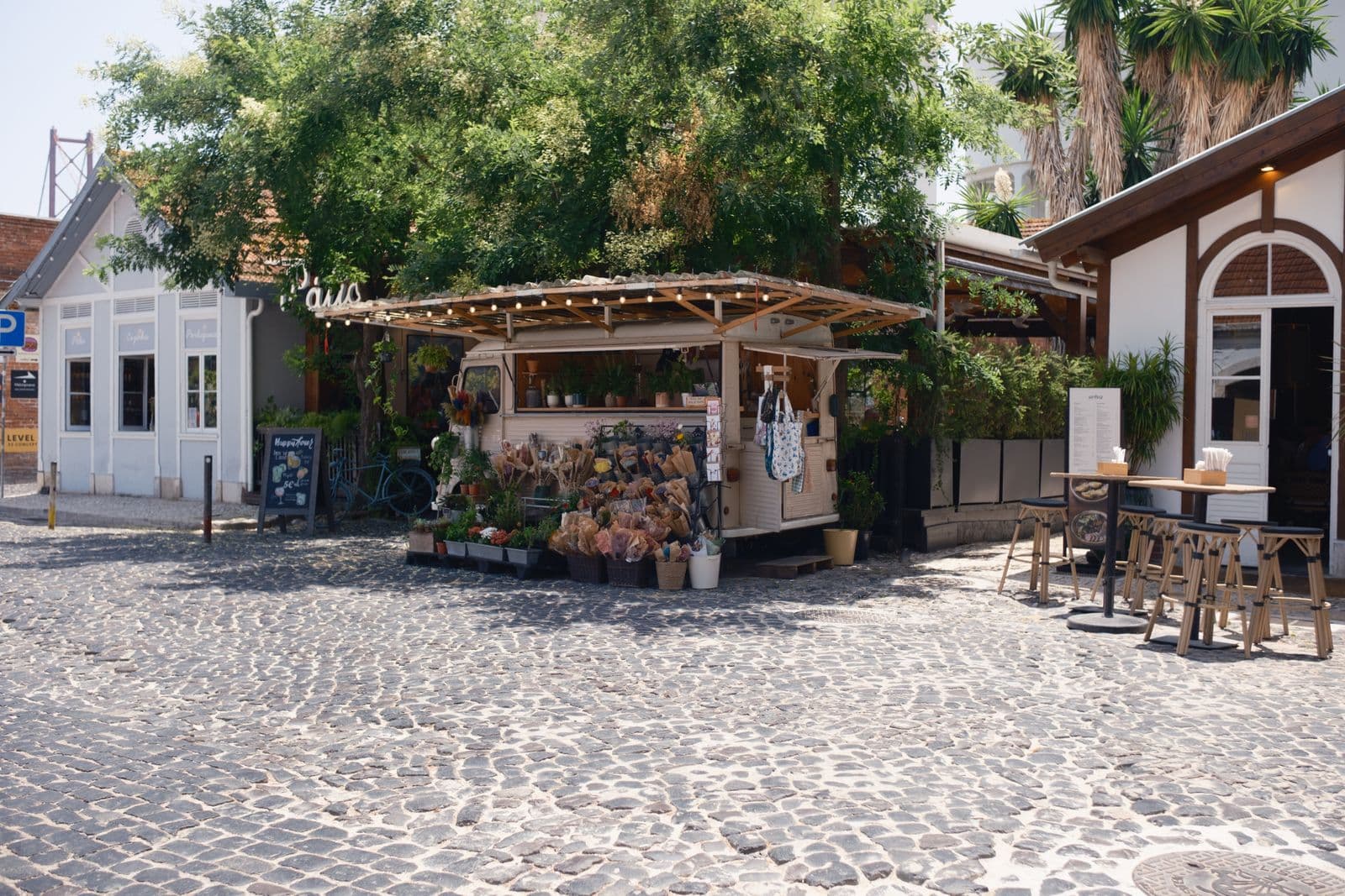 A flower stall built into an old cream-coloured van sits on a cobbled square in Lisbon, with bunches of dried flowers and potted plants in front and café tables to one side.
