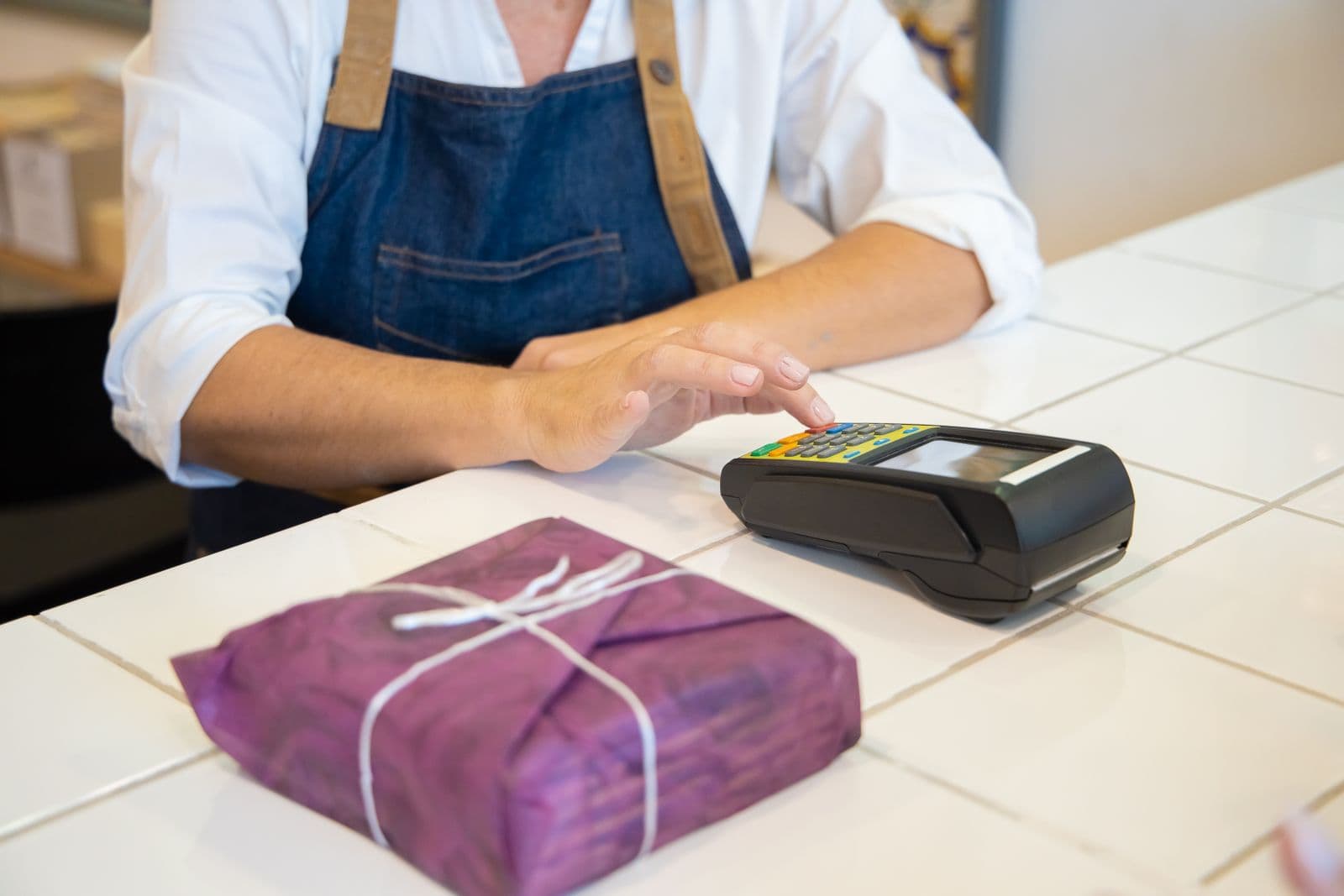 A shopkeeper in a denim apron keys an amount into a black card machine on a white tiled counter, beside a parcel wrapped in purple paper.