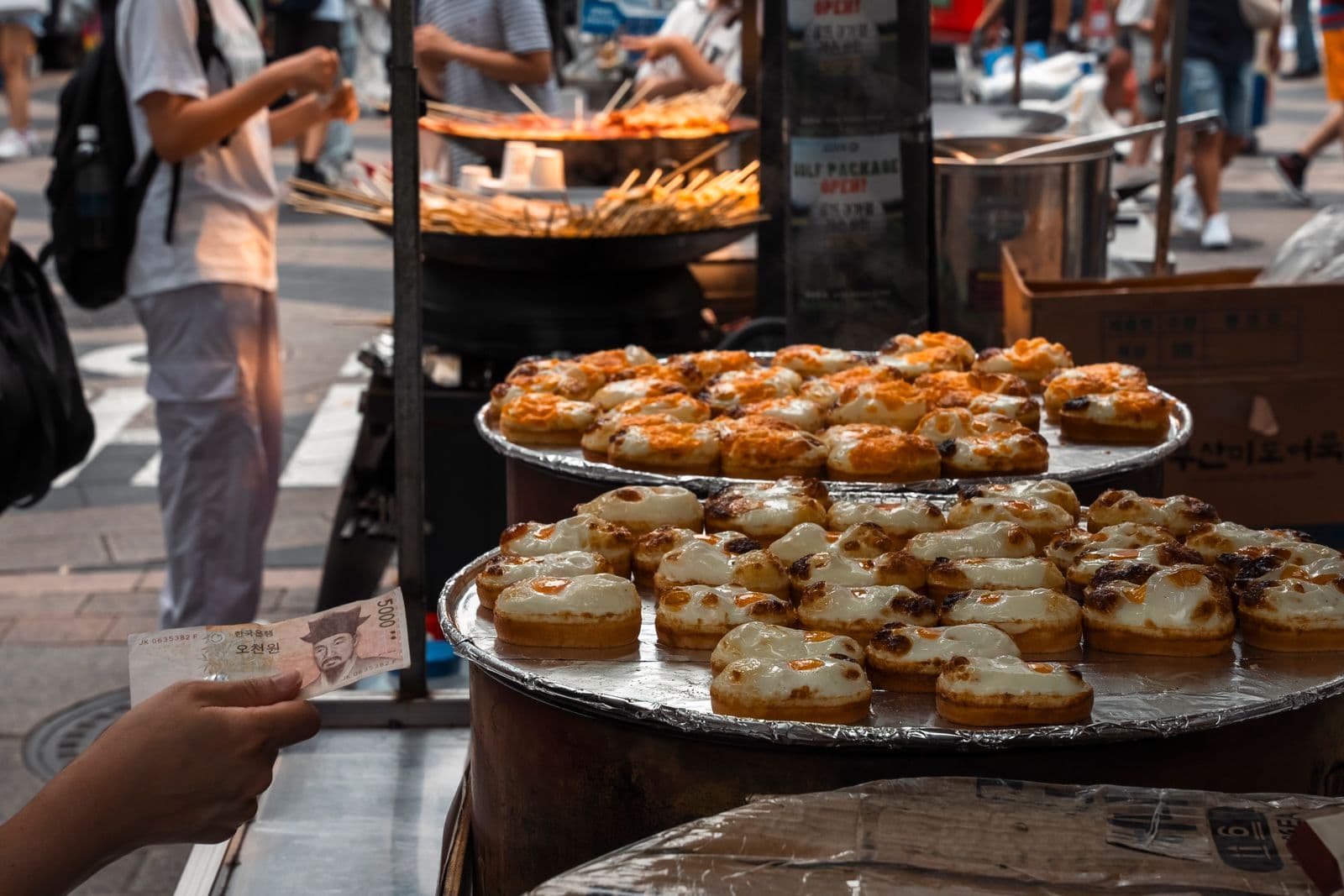 A customer's hand holds out a 5,000 won note at a Seoul street food stall, beside trays of Korean egg bread, with skewers cooking in a wide pan behind.