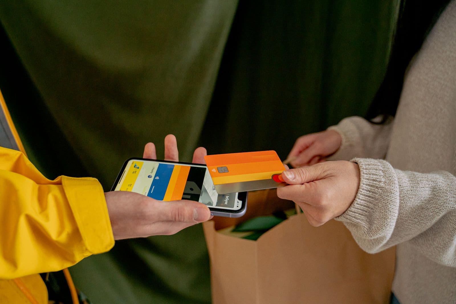 A customer in a knitted sweater holds an orange contactless card against the back of a smartphone held out by a person in a yellow jacket, with a paper shopping bag between them.