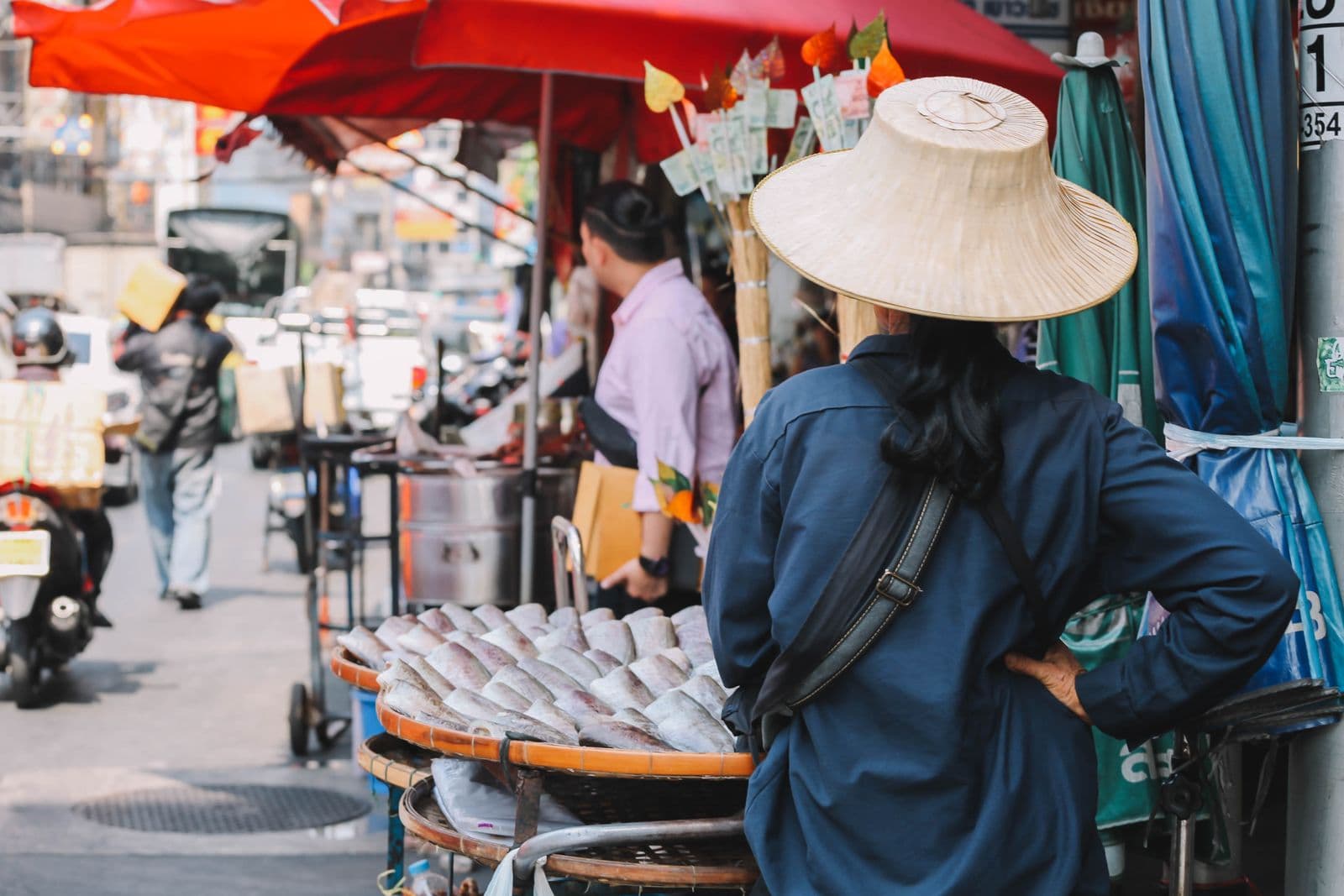 A street vendor in a wide woven straw hat and a navy work shirt stands with a hand on the hip beside round trays of dried fish on a Bangkok pavement, with red market umbrellas and a passing motorbike behind.