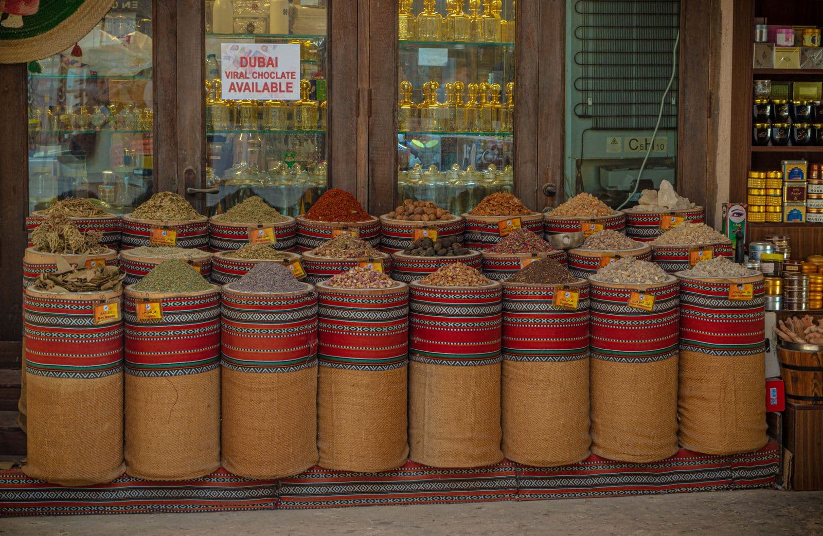 Woven drums of dried herbs, rosebuds, nuts and spices with handwritten labels stand in rows outside a shop in Dubai's spice souk, with perfume bottles on glass shelves behind.