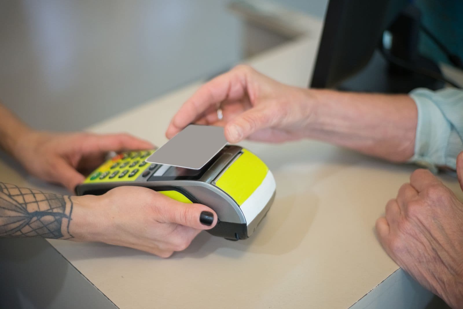 A customer holds a plain grey card against a lime-green card terminal that a shop assistant is holding out across a pale counter.